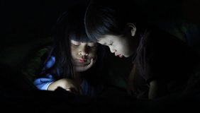 sister and brother using touch pad lying in bed at night. Bright screen lighting their faces