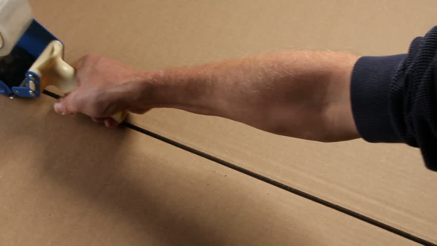 A hand pulls a tape gun across a corrugated cardboard box to tape up a seam.