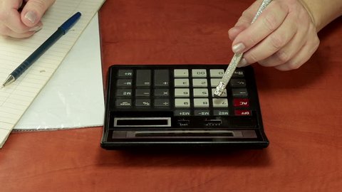 Woman left hand is pushing calculator buttons with pencil eraser and ready to record outcome by right hand