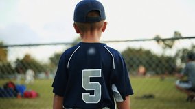Shot from behind of a cute kid in baseball uniform watching a practice behind a fence at baseball field