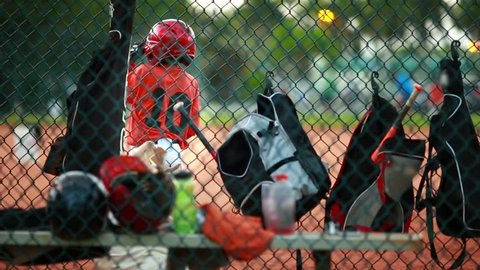 Shot from behind a fence at baseball field of baseball equipment such as bags and helmets and also of an unrecognizable kid swinging his bat during a game.