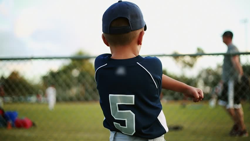 Shot from behind of a cute kid on his baseball uniform stretching his arms behind fence at baseball park. He is looking at a coach practicing with other kids. Unrecognizable kids and coach practicing
