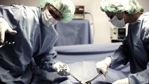 Two surgeons in full surgery garb, using surgical tools, operate on a patient in an operating room.