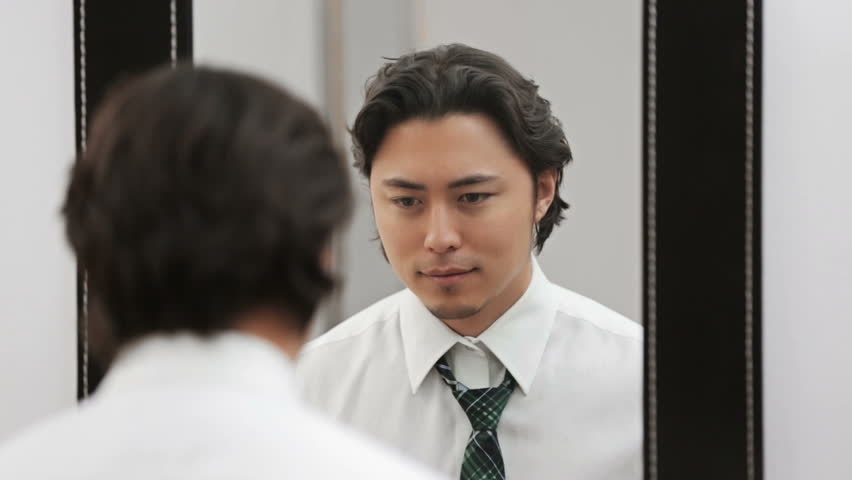 An attractive man wearing a shirt and tie, tying his tie in front of a mirror. Feeling confident and successful.