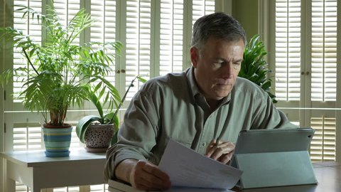 A mature serious man sitting at table in front of shuttered windows looking over financial statement and using an electronic tablet pc.