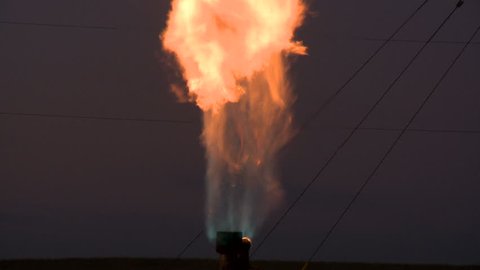 Tilt up on flames from a burning gas flare, at night.