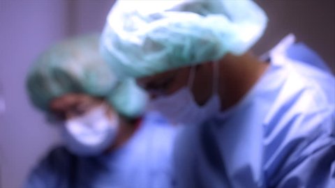 Camera moves from two out of focus surgeons working in the background to a surgical nurse in the foreground who stops and looks at the camera. Tinted for Affect