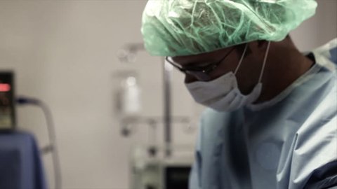 The camera pans and tilts from a male surgeon to a woman surgeon and a surgical nurse who stops assisting for a moment and stares into the camera.