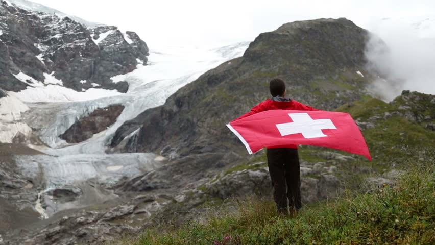 Switzerland hiker hiking cheering showing Swiss flag jumping in front of glacier. Happy Asian woman holding big red flag in nature.