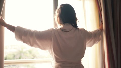 Woman in bathrobe unveil curtains in room in the morning 
