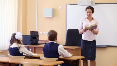 Teacher reads book near chalkboard and two pupils listen her in classroom at school