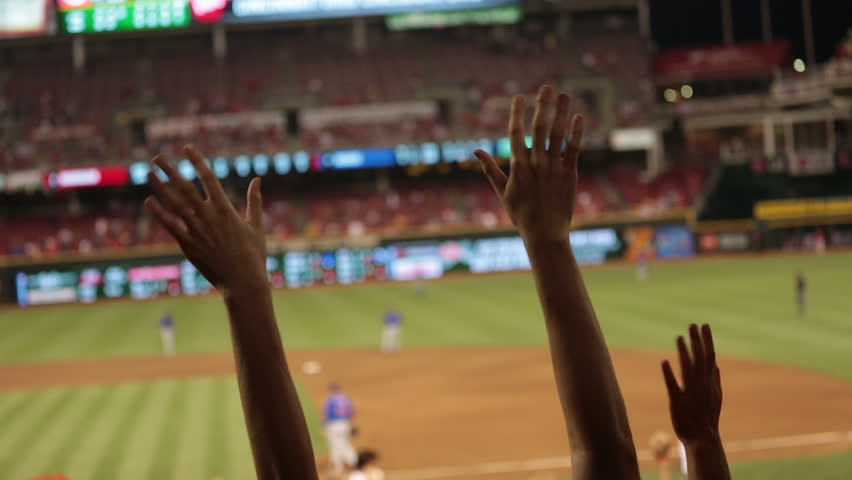 Two hands stick out and cheer during a baseball game. Gravel, green grass and baseball players soft focus in the background.