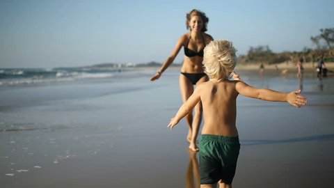 A little boy runs along the beach into her mothers arm's as the camera follows him. Slow motion 