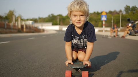Smiling little boy rides sitting on skateboard in slowmotion. Close up shot.