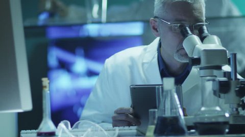  Scientist is Looking through Microscope and Using Tablet in Laboratory. Shot on RED Cinema Camera in 4K (UHD).