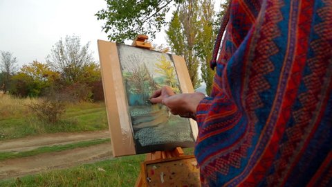 Portrait of a middle aged woman with long brown hair in warm clothes drawing a picture of an autumn landscape around her on the easel with a help of multi colored chalks. Camera is making a small zoom