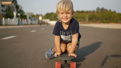 Happy little boy rides sitting on skateboard in slowmotion.