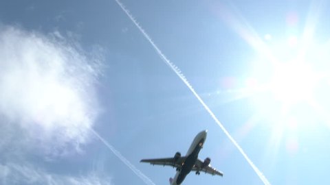 Commercial airliner flying overhead on sunny day with jet contrails lining the blue sky.