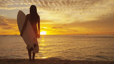 Young female surfer standing on beach sand under sunset sky holding surfboard watching ocean waves sunrise. Healthy lifestyle and sport concept. RED EPIC footage. Stockvideó