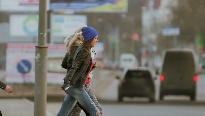 A group of young, active, cheerful people crossing the road. They run, laugh and rejoice in the background you can see the car traffic