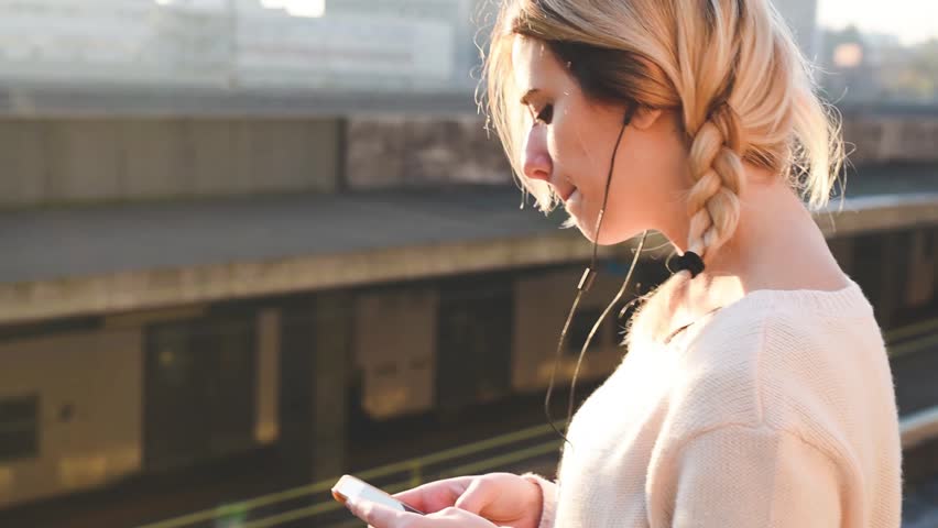 young beautiful blonde woman outdoor in the street of the city listening music and using smartphone with earphones in backlit sunset