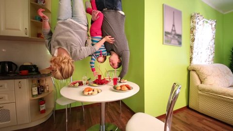 Parents and daughter upside down touch food on table in kitchen of inverted house