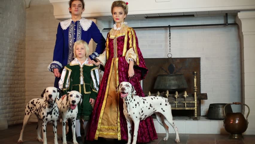 Father, mother and son in medieval costumes stand near fireplace with three dalmatians