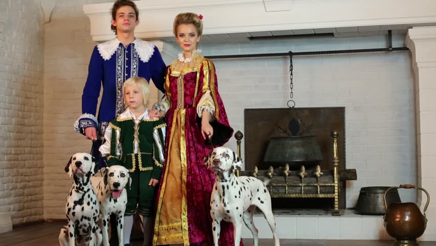 Father, mother and son in colorful medieval costume stand near fireplace with three dalmatians on leashes