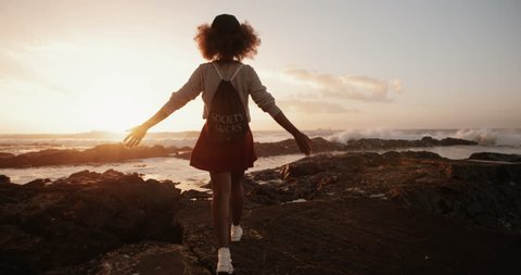 hipster girl walking and spinning on some rocks at the beach at sunset with sun flare, Slow Motion