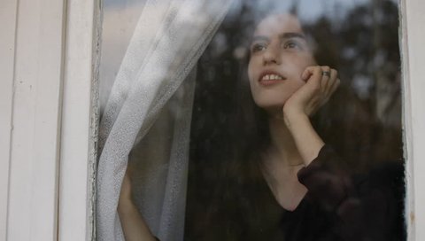 Brunette young adult woman opening drapes curtains, resting head in hand and looking through the window up to the sky. White painted wood frame. Medium shot. Daylight.