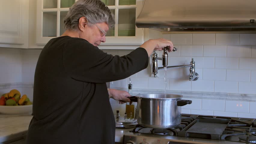 Mature woman in kitchen adds water to a cooking pot while preparing a meal in the stove.