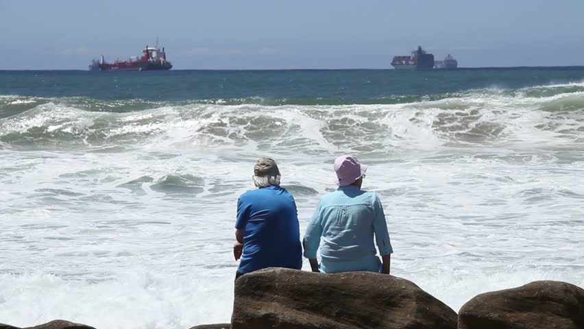 Retired couple watching large waves breaking with ships in the background.