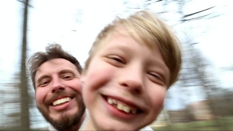 Father and son spinning around together while taking selfie in park. Point-of-view shot Funny family have fun outside. Happy dad and kid looking at camera. Smiling laughing man and child. Spring park