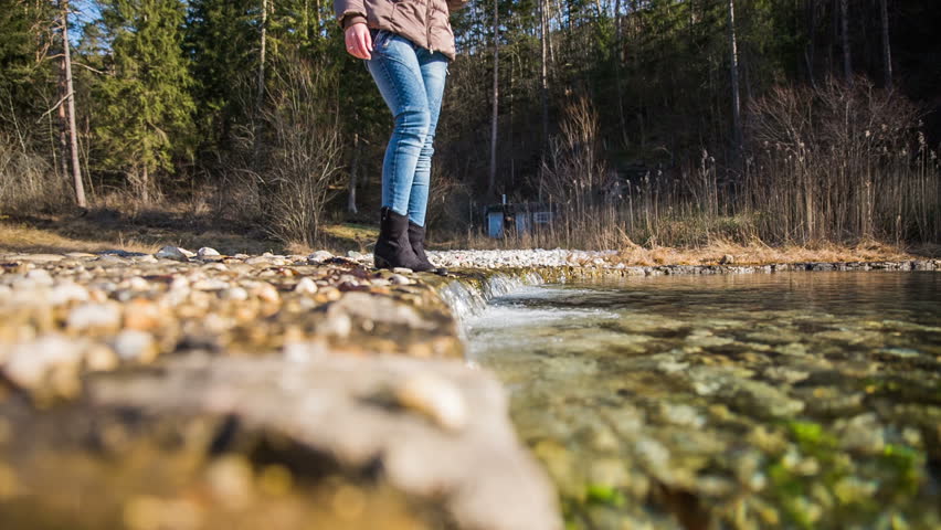 Woman soak hand in small river in nature. Female person in warm jacket standing over a small clean river running in to lake and putting hand under waterfall.