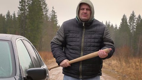 Aggressive man with a baseball bat near car in rainy day
