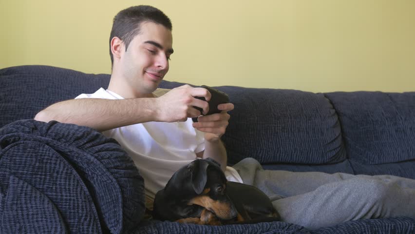 Caucasian attractive young adult laying on the livingroom couch and texting on his smartphone. A pinscher pet dog is sitting with him keeping him company.