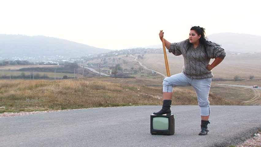 Slow motion - shot of a mad plus-sized black-haired woman in blue jeans and grey sweater with swinging a wooden club in her hands standing on the road near TV-set screaming and pointing at something