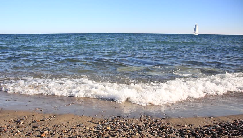 Sailboat on blue water, waves washing upon sand beach, clear blue sky