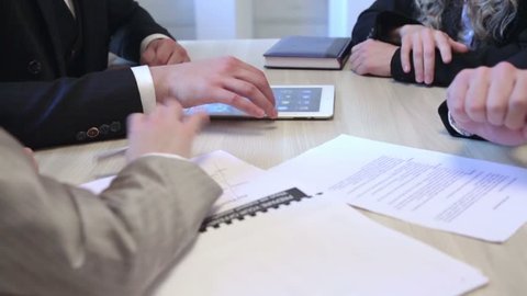 Partners are meeting in the office. Businessmen negotiate behind the table. They are discussing and doing handshake. The tablet and documents are on the table.