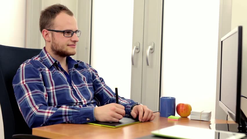 Young, handsome man in glasess, sitting behind the desk and using computer. He is surprised when he gets a lot of paper work to do. 