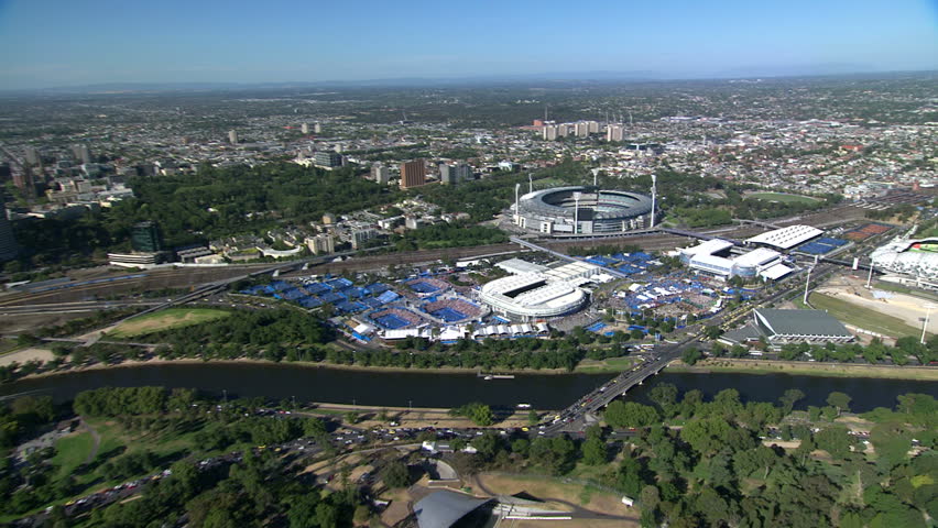 A wide sweeping aerial shot of Melbourne City, Victoria, Australia, from a helicopter, across the green botanical gardens and Yarra River towards the tennis stadiums and MCG sports ground.