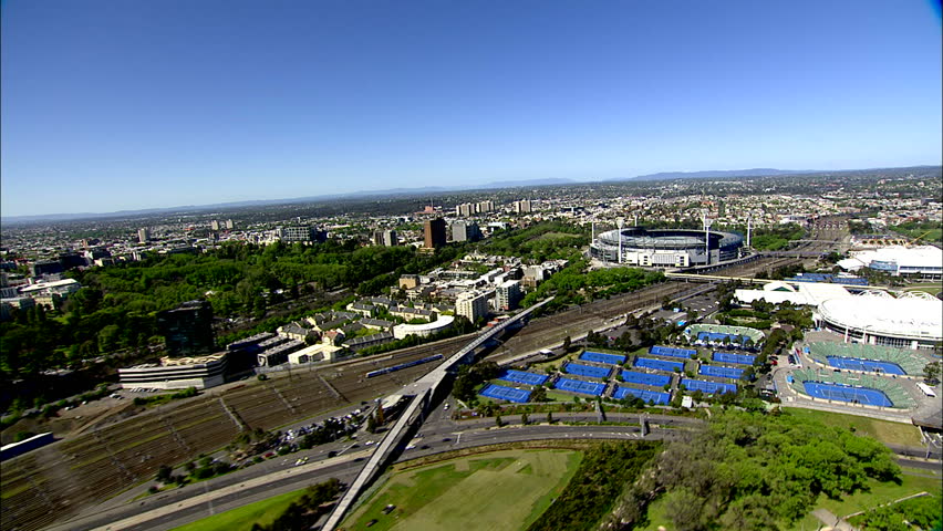 A wide sweeping aerial shot of Melbourne City, Victoria, Australia, from a helicopter, across the green botanical gardens towards the tennis stadiums and MCG sports ground.