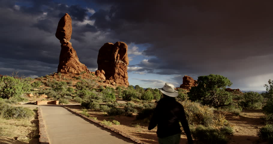 Arches National Park, Moab Utah, USA. Female hiker passing camera and walking on path towards Balanced Rock at golden hour sunset with dramatic dark clouds in the distance.