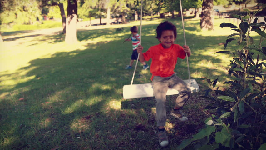 Happy little boy on a swing in the park, friend plays in the background