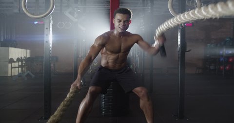 Medium shot slow mo zoom-in, zoom-out young man exercising arms and shoulders with rope in gym