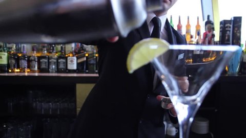 Barman makes a cocktail and than he pours it the cocktail glass