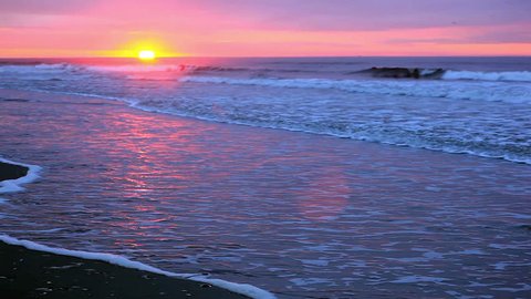 Beach sunrise.  Sun is at the horizon with waves and shore showing.  Pretty pinks, grays and purples in the sky, sea and shore and very nice reflections.