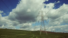 Real time clip of high voltage power lines and tall electricity pylons extending to the horizon against the sky and clouds,in a vast green field in the countryside.