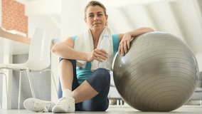 Mature woman drinking water, sitting by fitness ball