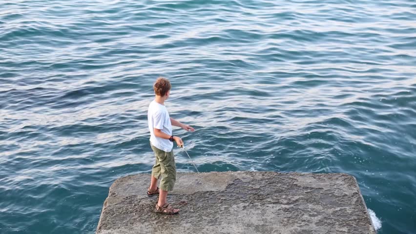 Boy with fishing rod fishes in sea and stands on concrete place at summer